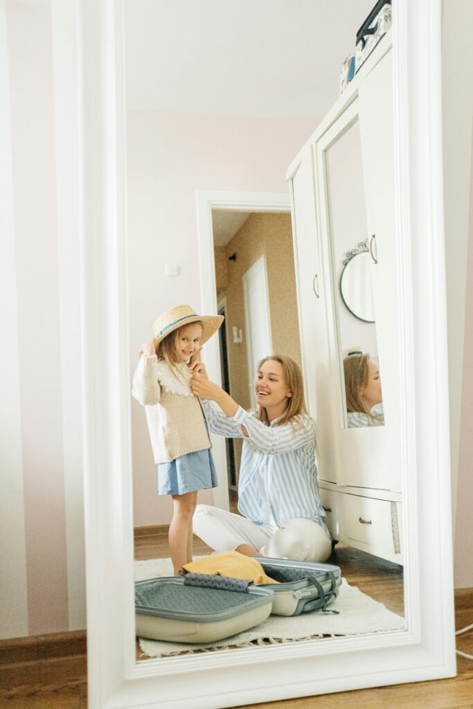 A mother assists her young daughter in trying on a hat while packing for a vacation indoors.